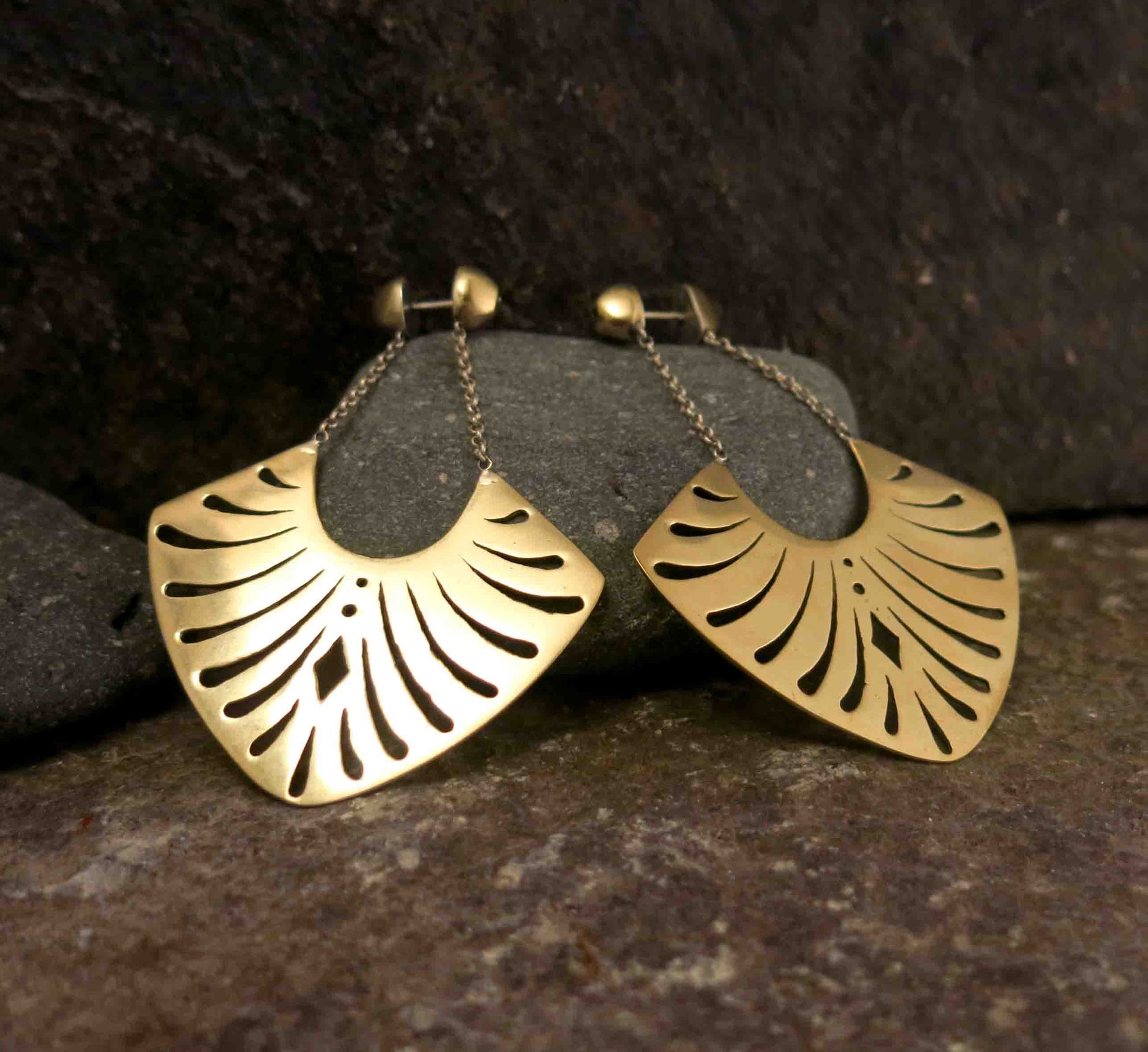 A pair of brass earrings featuring a fan-like shape with geometric cut-outs, hanging from silver chains attached to rounded stud tops. Photographed on a stone. 
