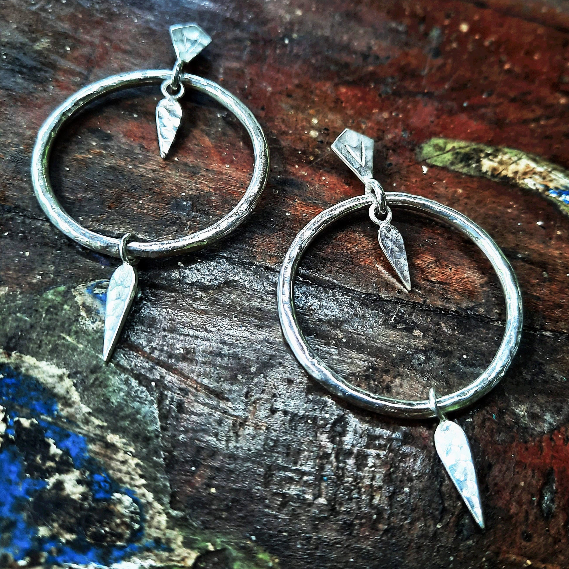 Sterling silver-plated circular earrings with a hammered texture, adorned with two elongated pendants suspended from the hoop. The pendants add movement and an artisanal touch, paired with silver ear hooks.
