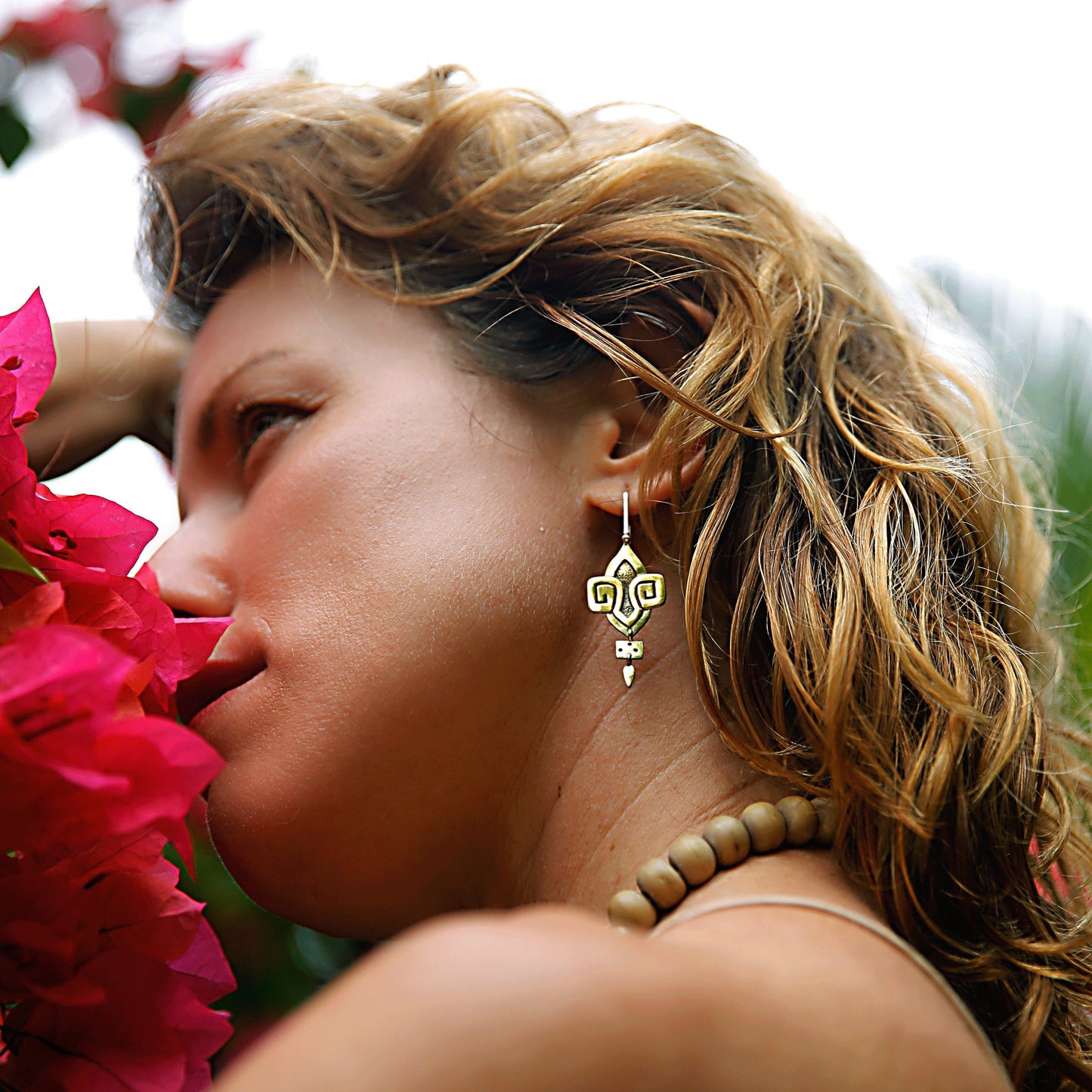 Model smelling red flowers and wearing the Hadza earrings in warm, golden Italian brass with geometric tribal-inspired shapes and hammered textures.